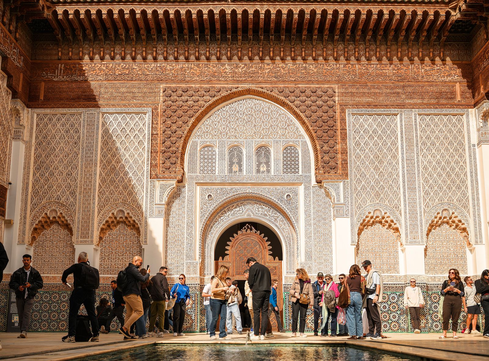 Un groupe de touristes explore l'architecture complexe de la médersa Ben Youssef sous le soleil de Marrakech.